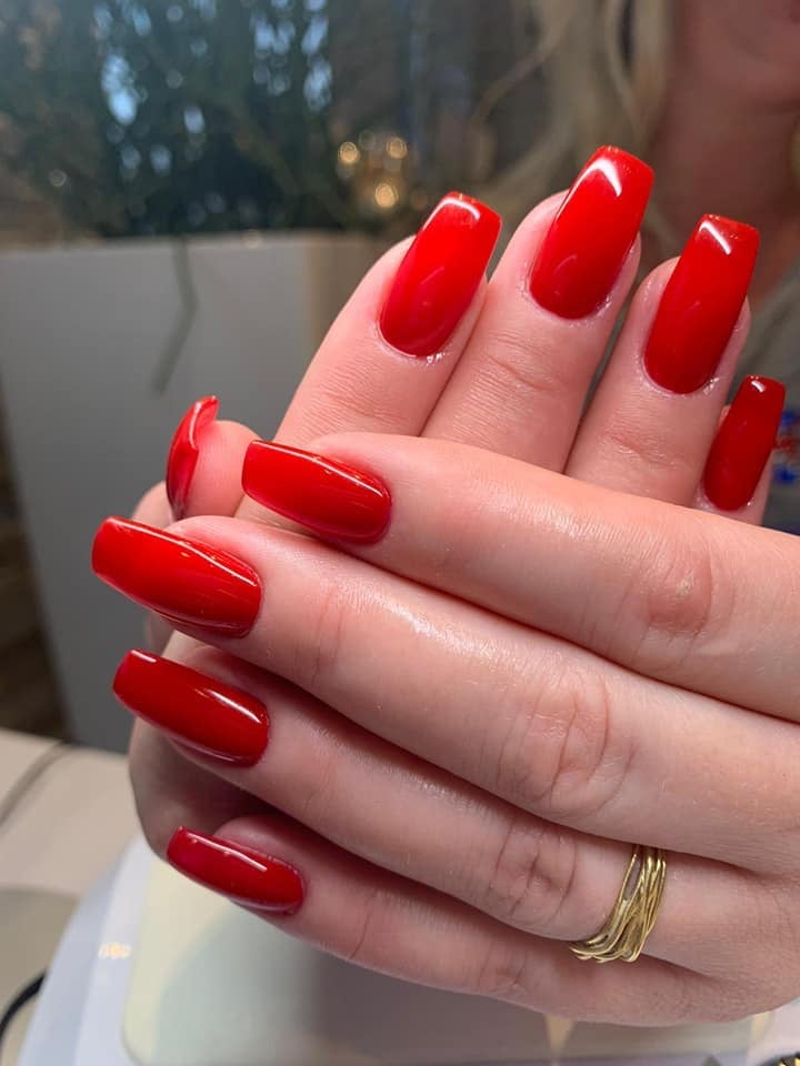 Close-up of manicured hands with glossy red gel nails and gold rings against blurred background